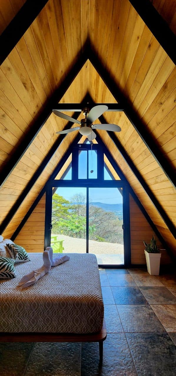 A-frame bedroom with triangular window framing mountain views and bed with geometric bedding, an upscale comfort